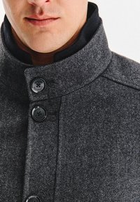 Szara marynarka w jodełkę z wysokim kołnierzem, wyposażona w czarne guziki i kontrastującą podszewkę wewnętrzną. Teksturowana tkanina podkreśla wizualną głębię.