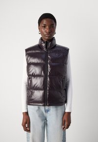 Tamno ljubičasti, puferasti prsluk bez rukava s visokim ovratnikom, zatvaranjem na patentni zatvarač sprijeda i bočnim džepovima. Glatki, reflektirajući materijal s suptilnim šavovima.