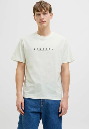 Joven vestido con una camiseta blanca con el texto "JJREBEL" en el frente y jeans azules, posando frente a un fondo gris claro liso.