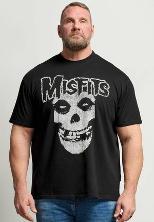 Muskuløs mand med kort hår og skæg bærer en sort Misfits band T-shirt med en stor hvid kraniegrafik foran.