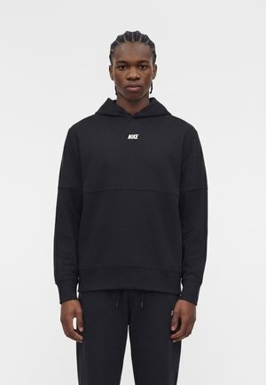 Jeune homme aux cheveux tressés portant un sweat à capuche noir Nike et un pantalon noir, debout devant un fond clair uni.