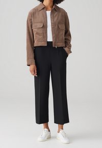 Veste cropped en daim marron avec deux poches avant, associée à un pantalon large noir et des baskets blanches. Design texturé et contemporain.