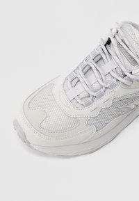 Hästi lähedalt vaadatuna helehall vabaajajalanõu, millel on võrkmaterjalist osad, tugevdatud varvas ning valged paelad valgel taustal.