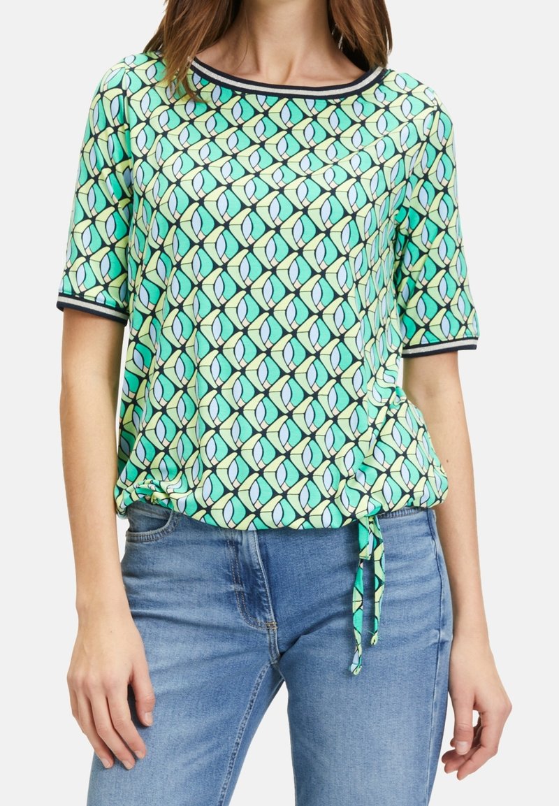 Femme portant un haut à manches courtes, à motifs verts, bleus et jaunes avec une bordure noire et un nœud devant, associé à un jean bleu.
