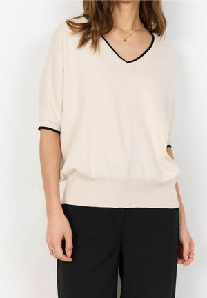 Femme portant un pull crème à manches courtes en V avec un bord noir, assorti à un pantalon noir et des colliers en or superposés.