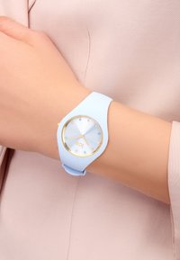 Montre en silicone bleu clair avec des accents dorés, cadran rond et index horaires en cristal. Le bracelet est lisse, flexible et s'ajuste parfaitement au poignet.