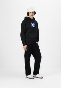 Black hooded sweatshirt with a blue graphic logo, front pocket, paired with black pants and white sneakers, completed by a light cap.