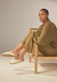 Vita espadrillos med flätad jutekant, parat med en lös olivgrön linneoutfit, sittande på en stol av naturligt trä med vävd sits.