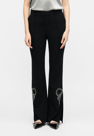 Γυναίκα που φοράει μαύρο παντελόνι με φαρδύ πόδι, λευκή κεντημένη ανθοδέσμη κοντά στο τελείωμα και πλαϊνές σχισμές, συνδυασμένο με μαύρα μυτερά παπούτσια.