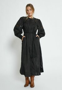 Black long-sleeve dress with textured fabric, puffed sleeves, waist tie, and side pockets, paired with brown ankle boots.