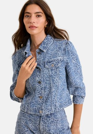 Noor naine pikkade pruunide juustega, kandes sinist teksatakki ja sobivat pükst, millel on valged lillemustrid.