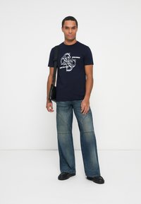 Hombre de pie vistiendo una camiseta gráfica azul marino, pantalones vaqueros de pierna ancha azules, zapatos negros y un bolso negro de hombro contra un fondo blanco liso.