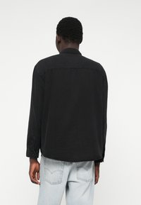 Juodų džinsų marškiniai su ilgomis rankovėmis, mygtukinėmis apykaklėmis ir tiesiu apačiu. Dėvimi su šviesiai mėlynomis džinsinėmis kelnėmis, demonstruojantys kasdienį stilių.