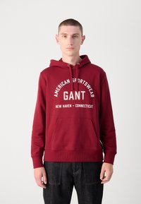 Burgundi kapucnis pulóver elülső zsebbel, húzózsinórral és fehér szöveggel, ami azt írja: "American Sportswear GANT New Haven - Connecticut." Pamutkeverék anyag.