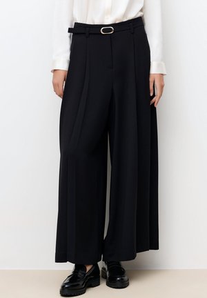 Kobieta nosząca czarne spodnie z wysokim stanem i szerokimi nogawkami z cienkim czarnym paskiem oraz czarne mokasyny, w połączeniu z białą bluzką z długim rękawem.