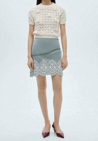 Haut en maille crème à manches courtes, associé à une jupe bleu clair ornée d'une bordure en dentelle blanche délicate et d'un ourlet festonné. Talons hauts rouges.