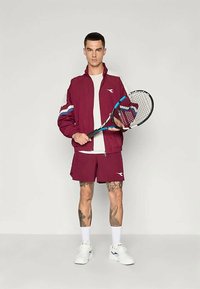 Μαρούλι αθλητικό μπουφάν με μπλε και άσπρες ρίγες, συνδυασμένο με ταιριαστά σόρτς. Κρατάει μια ρακέτα τένις, τα παπούτσια είναι λευκά και οι κάλτσες είναι ορατές μέχρι τον αστράγαλο.