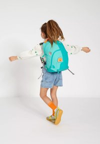 Bambino con lunghi capelli legati in una coda di cavallo, che indossa una blusa con stampa di stelle, pantaloni corti di jeans, calzini arancioni e scarpe da ginnastica, con le braccia aperte e uno zaino turchese con un patch di cartone animato.