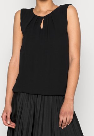 Woman wearing a sleeveless black blouse with a small keyhole neckline and a black pleated skirt.
