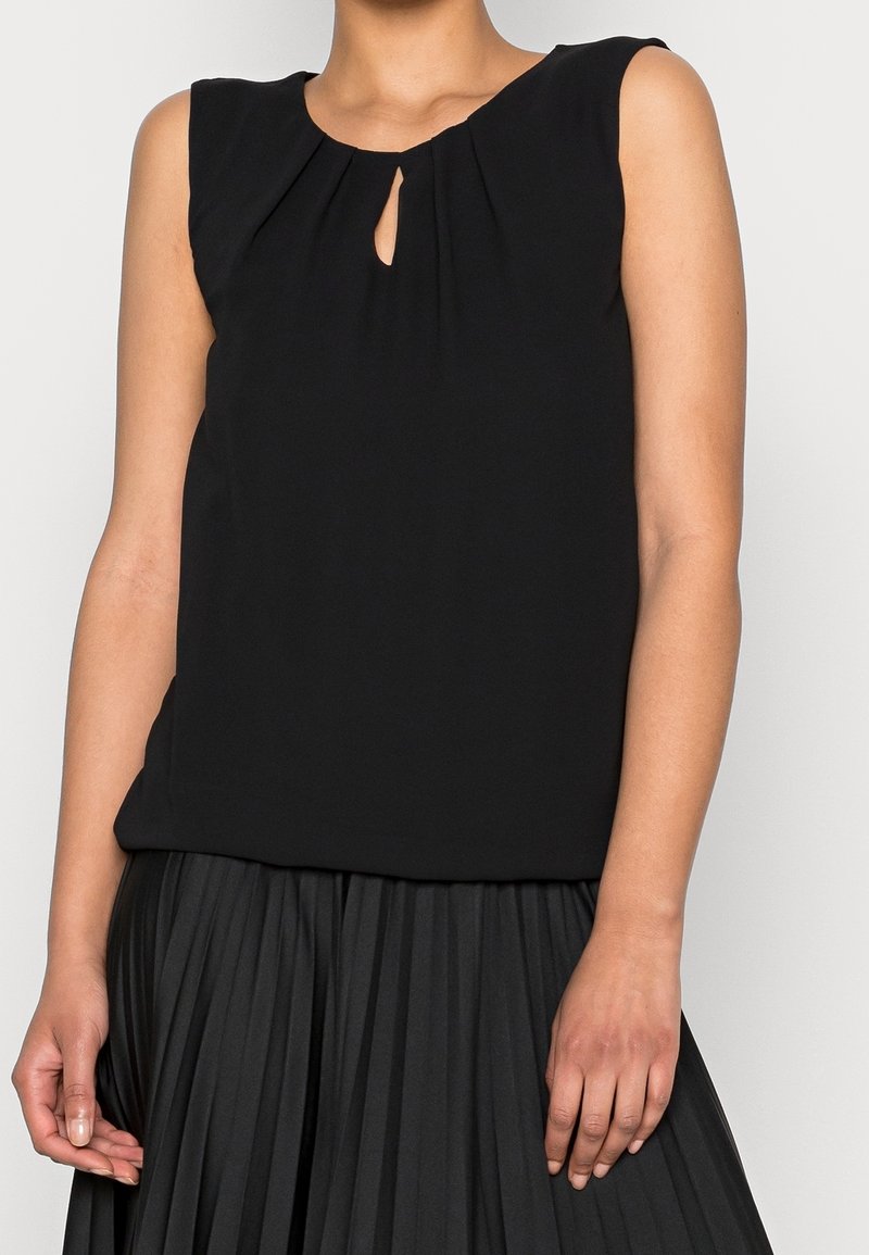 Femme portant un chemisier noir sans manches avec une petite ouverture en forme de goutte au niveau du col et une jupe plissée noire.