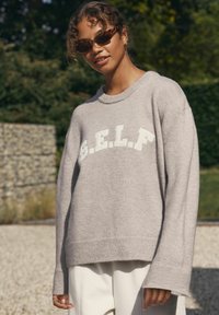 Maglione grigio a maglia con vestibilità ampia, scollatura rotonda e design "S.E.L.F." in bianco sulla parte anteriore. Texture morbida e maniche lunghe.