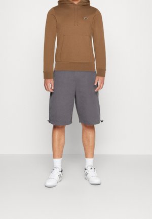 Homme portant un sweat à capuche marron, un short gris jusqu'aux genoux, des chaussettes blanches et des baskets blanches, debout devant un fond uni.