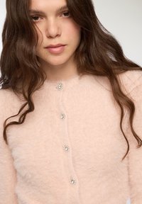 Cardigan fluffy rosa chiaro con una texture morbida, scollatura rotonda e cinque chiusure decorative con bottoni dotati di dettagli simili a gemme.