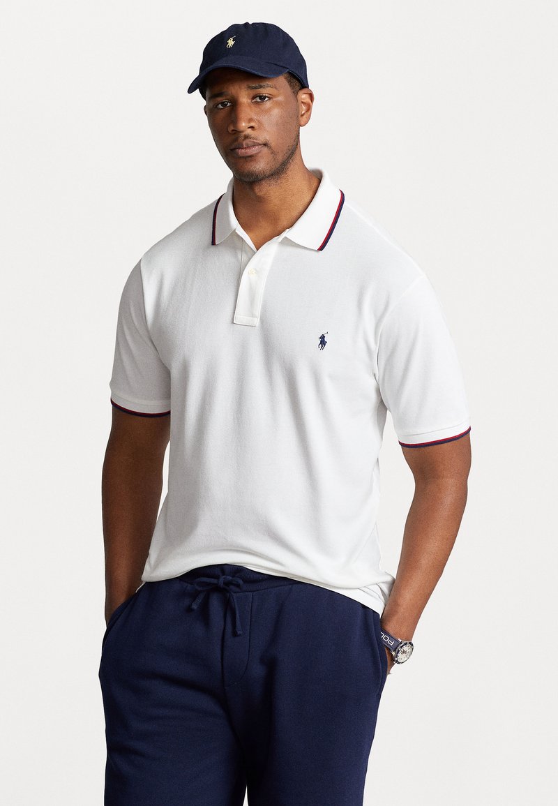 Chemise polo blanche avec un plastron à deux boutons et des accents de col rouge et bleu, associée à un pantalon navy à cordon de serrage et une casquette navy.