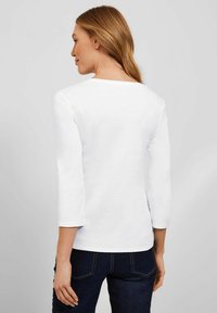 Witte, langetermijn overhemd met een ronde halslijn, gemaakt van gladde stof. Het heeft driekwartmouwen en een getailleerd ontwerp, gecombineerd met donkerblauwe jeans.
