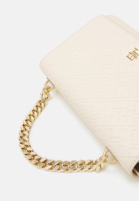 Cremfarbige Handtasche mit geprägtem geometrischem Muster, ausgestattet mit einem goldenen Kettenriemen und einem Logoakzent auf der Vorderseite.