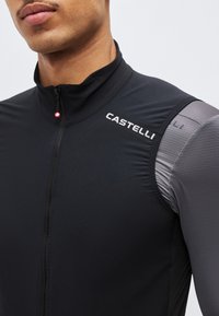 Κοντινό πλάνο ενός ατόμου που φοράει μαύρο αμάνικο ποδηλατικό γιλέκο Castelli με φερμουάρ πάνω από γκρι μακρυμάνικη υφασμάτινη μπλούζα.