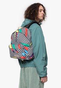Checkerboard-patterned backpack with multicolored geometric accents. Features fabric straps and a front pocket. Compact, rounded design.