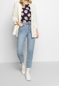 Bílý blazer přes tmavý květinový top s červenými a bílými sedmikráskami, spárovaný se světle modrými rovnými džíny a bílými teniskami.