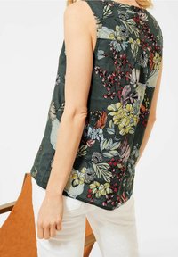 Green floral sleeveless top featuring colorful embroidered patterns, round neckline, and relaxed fit; paired with white pants.
