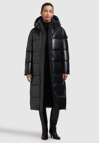Dlouhý černý lesklý péřový kabát s kapucí, s prošívaným designem, předním zipem a bočními kapsami. Nositý přes černý outfit a elegantní boty.