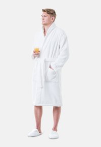 Witte badjas van badstof met een ceintuur in de taille, zijzakken, gedragen met bijpassende witte slippers. Het model houdt een glas sinaasappelsap vast.