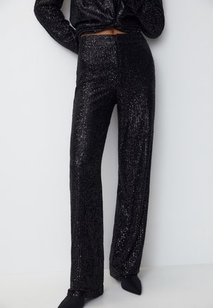 Woman wearing black sequined high-waisted wide-leg pants and matching cropped top, standing against a plain white wall.