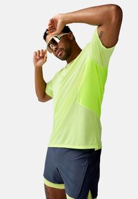 Neon geltonas, trumpomis rankovėmis sportinis marškinėlis su tekstūruotu audiniu ir kišene. Sujungtas su tamsiai pilkomis šortais, turinčiais šviesesnį apdailą.