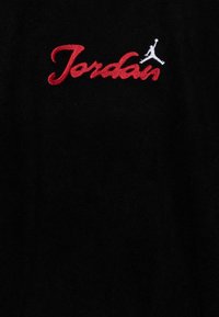 Melnas auduma plāksne ar izšūtu "Jordan" sarkaniem burtiem un baltu siluetu logotipu. Dizains ir centrēts un izteiksmīgs uz materiāla.