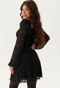 Svart spetsklänning med långa, utsvängda ärmar, texturerad yta och en åtsittande livdel. Kjolen har en utsvängd silhuett och slutar strax ovanför knäet.