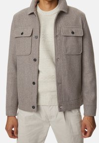 Grå uldblanding jakke med to brystlommer, sorte knapper og krave, lagdelt over en tekstureret cremefarvet sweater. Velegnet til køligt vejr.