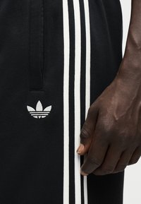 Μαύρο παντελόνι Adidas με τρεις λευκές κάθετες ρίγες και λογότυπο κοντά στην τσέπη, μαζί με κοντινή εικόνα ενός σκουρόχρωμου χεριού.