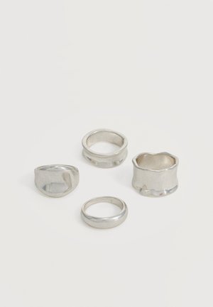 Quatre bagues en argent de largeurs variées et aux formes uniques, disposées librement sur un fond clair uni.