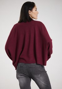 Burgunderfarbener Oversized-Pullover mit geripptem Saum, weicher Textur und Fledermausärmeln, kombiniert mit eng anliegenden grauen Jeans mit Gesäßtaschen.