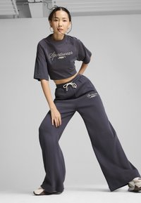 T-shirt cropped grigio scuro con testo "Sportswear by PUMA", abbinato a pantaloni jogger a gamba larga coordinati e sneakers di colore chiaro. Texture del tessuto casual.