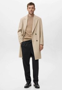 Abrigo beige de doble botón, mezcla de lana, con hombros estructurados; suéter marrón claro, pantalones negros ajustados; zapatos negros con cordones.