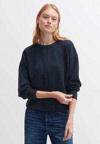 Felpa blu navy con collo a giro, spalle scese e polsini a coste. Tessuto morbido con vestibilità comoda, indossata sopra jeans blu.