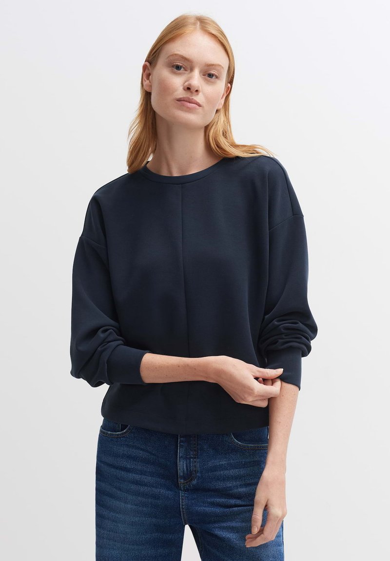 Felpa blu navy con collo a giro, spalle scese e polsini a coste. Tessuto morbido con vestibilità comoda, indossata sopra jeans blu.