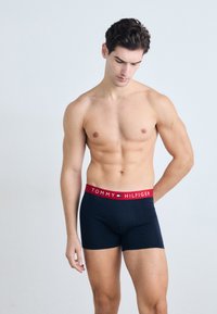 Boxeri navy cu talie roșie, având inscripția "TOMMY HILFIGER" în alb. Materialul este neted și elastic, punând în evidență un design minimalist.
