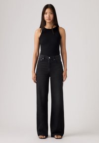 Νεαρή γυναίκα με μακριά μαύρα μαλλιά, φοράει μαύρο αμάνικο τοπ, μαύρα τζιν παντελόνια με ψηλή μέση και φαρδιά μπατζάκια, και μαύρες σαγιονάρες, στέκεται μπροστά σε γκρι φόντο.